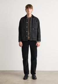 Black denim jacket with button closure, two front pockets, worn over a grey sweater, paired with black pants and black shoes.