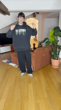 Hoodie preto com texto branco a escorrer e calças de fato pretas a condizer. Ténis prateados metálicos. Ambiente interior com uma planta e um sofá castanho.