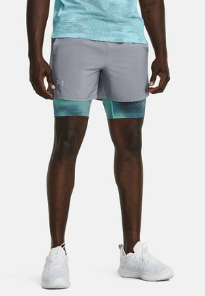 Mann trägt graue Under Armour Laufshorts über blau gemusterten Kompressionsshorts und weißen Laufschuhen, steht aufrecht.