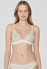 Krajková podprsenka v světle béžové barvě s trojúhelníkovým tvarem, vroubkovanými okraji a širokým elastickým pásem, s tenkými nastavitelnými ramínky.
