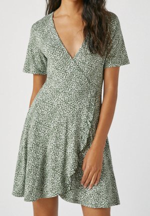 Frau trägt ein kurzes, grünes Wickelkleid mit kleinem weißen Blumenmuster, V-Ausschnitt und gerafftem Saum, steht vor einfachem Hintergrund.