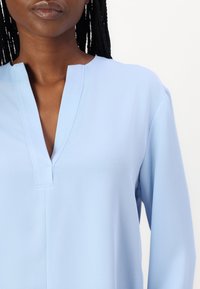 Blusa azzurra chiara con scollatura a V, maniche lunghe e texture liscia. Presenta cuciture minime e una vestibilità morbida.