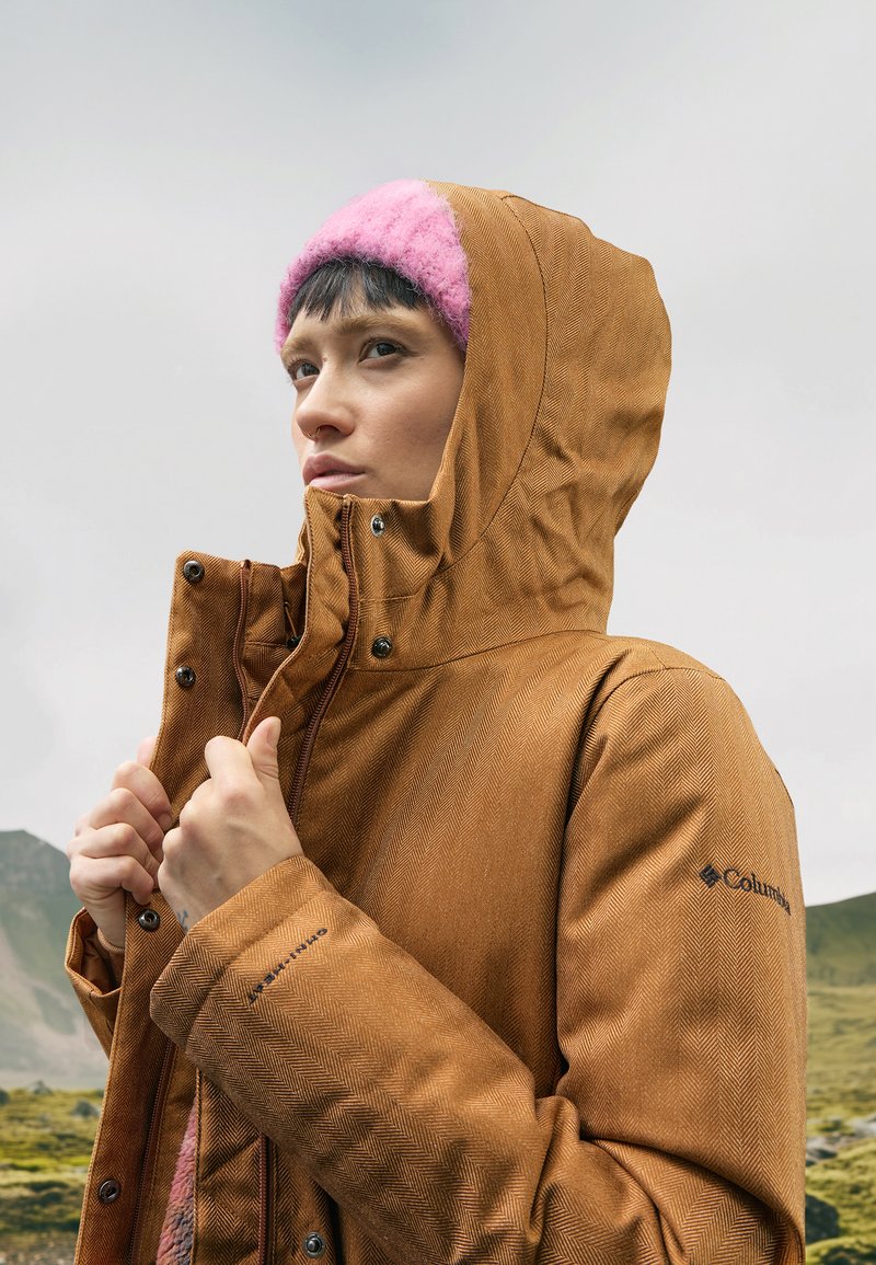 Braune Jacke im Fischgrätmuster für den Außenbereich mit hohem Kragen, verstellbarem Kapuzen und schwarzem Logo am Ärmel. Getragen mit einer rosa Strickmütze.