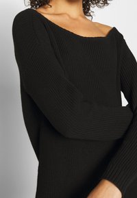 Černý pletený svetr s širokým off-shoulder výstřihem, žebrovanou texturou a dlouhými rukávy se staženými manžetami.