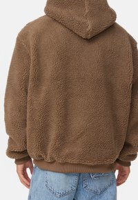 Brun sherpa fleece hoodie som bärs av en person som vänder bort, ihop med ljusblå denimjeans, som visar baksidans detaljer och textur.