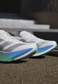 Een paar witte hardloopschoenen met blauwe en groene zolen die rusten op een gestructureerde ondergrond nabij betonnen trappen.