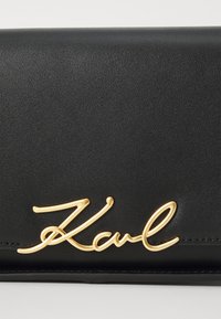 Czarny portfel z ekoskóry z złotym, kursywowym logo "Karl". Gładka faktura z zapięciem na zgięcie i bez widocznych detali metalowych.