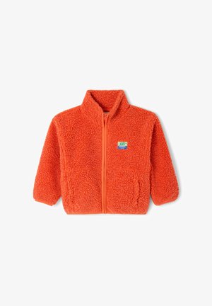 Lokkis juustega laps kannab üle suure oranži flisitagi, oranži dressipluusi, lilla lühikesi pükse ja beeže libisevaid jalanõusid, seisab valge seina taustal.