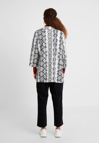 Zwart-witte jas met slangenprint en driekwart mouwen, gecombineerd met zwarte taps toelopende broek en witte sneakers.