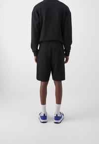 Homme se tenant de dos, portant un haut à manches longues noir, un short noir avec une poche arrière, des chaussettes blanches et des baskets blanches avec des accents bleus.