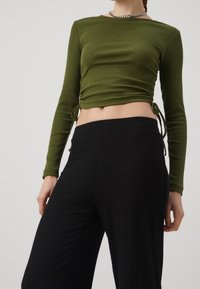 Vrouw in een olijfgroene gerimpelde langslap crop top en hoge zwarte ribgebreide wijde broek, met een zilveren ketting.
