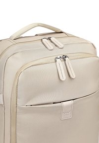 Krémový látkový batoh s dvojitými zipovými přihrádky, horním uchem a přední kapsou s logem. Hladká textura a pevný tvar.