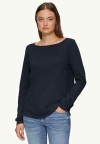 Navyblauwe longsleeve shirt met een ronde hals en zachte, gestructureerde stof. Gepaarde met lichtblauwe jeans, handen in de zakken.