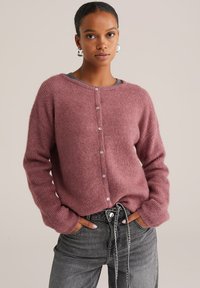 Femme portant un cardigan en tricot rose poussiéreux avec des boutons et des manches longues, légèrement rentré dans un jean noir délavé taille haute à cordon.