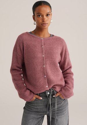 Femme portant un cardigan en tricot rose poussiéreux avec des boutons et des manches longues, légèrement rentré dans un jean noir délavé taille haute à cordon.