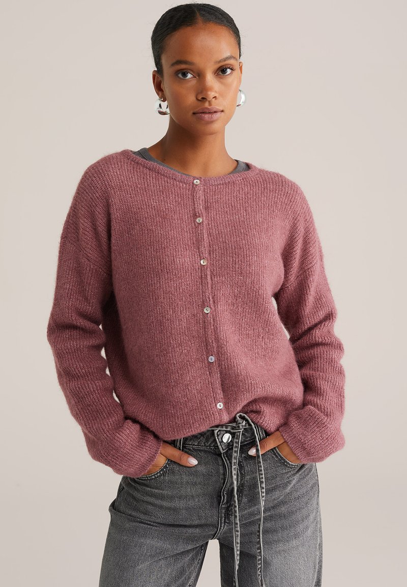 Femme portant un cardigan en tricot rose poussiéreux avec des boutons et des manches longues, légèrement rentré dans un jean noir délavé taille haute à cordon.