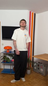 Jaunas vyras su tatuiruotėmis, dėvintis baltą marškinėlį ir juodas kelnes, stovi kambaryje su retro dryžuotu sienu, oranžine lempa ir augalais.