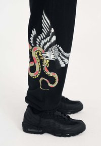 Svarta byxor har ett detaljerat tryck av en örn och en orm i livliga färger. Svarta Nike Air Max 95-sneakers bärs.