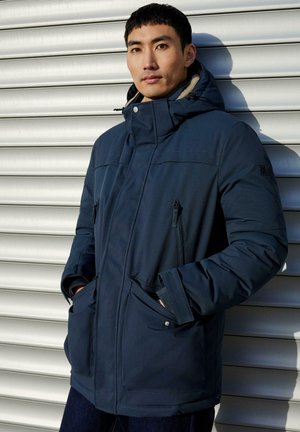 Man in een marineblauwe gewatteerde winterjas met capuchon, staand tegen een lichtgrijze metalen rolluik achtergrond, met handen in de zakken.