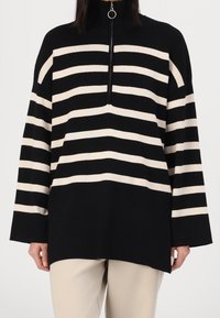Jersey de rayas negras y crema con un cuello alto, que presenta un cierre de media cremallera y mangas anchas. Hecho de un tejido de punto suave.