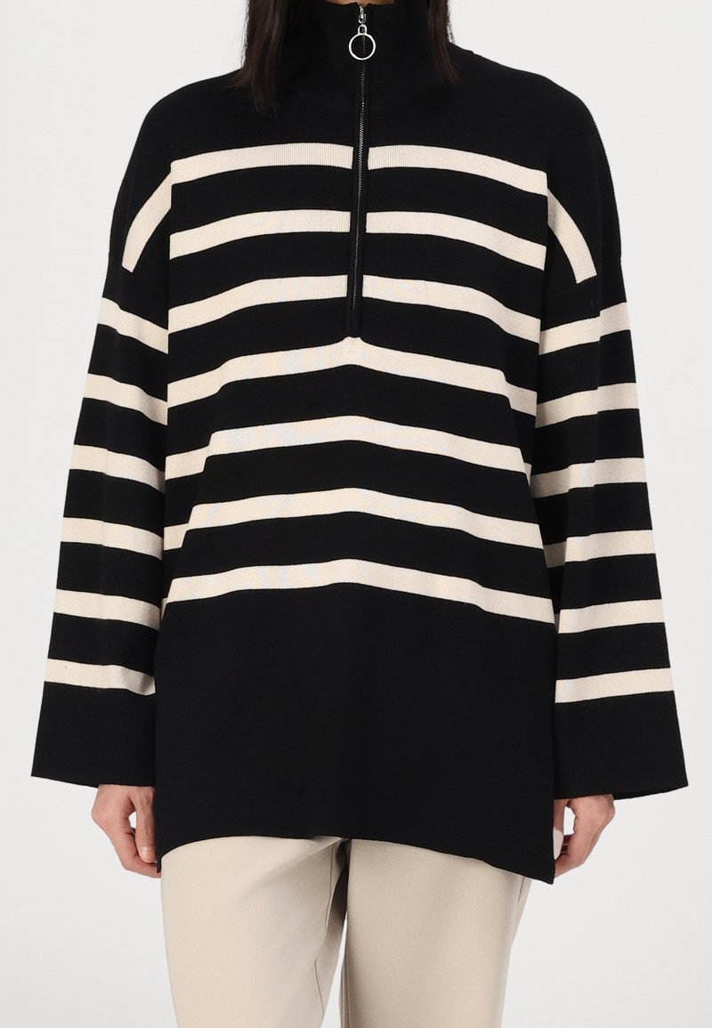 Jersey de rayas negras y crema con un cuello alto, que presenta un cierre de media cremallera y mangas anchas. Hecho de un tejido de punto suave.