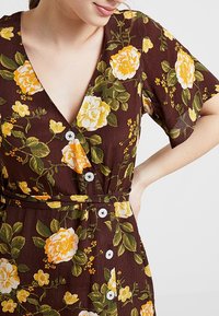 Robe à motif floral marron avec des roses jaunes et blanches, manches courtes, décolleté en V et détails en boutons, accompagnée d'une ceinture en tissu.