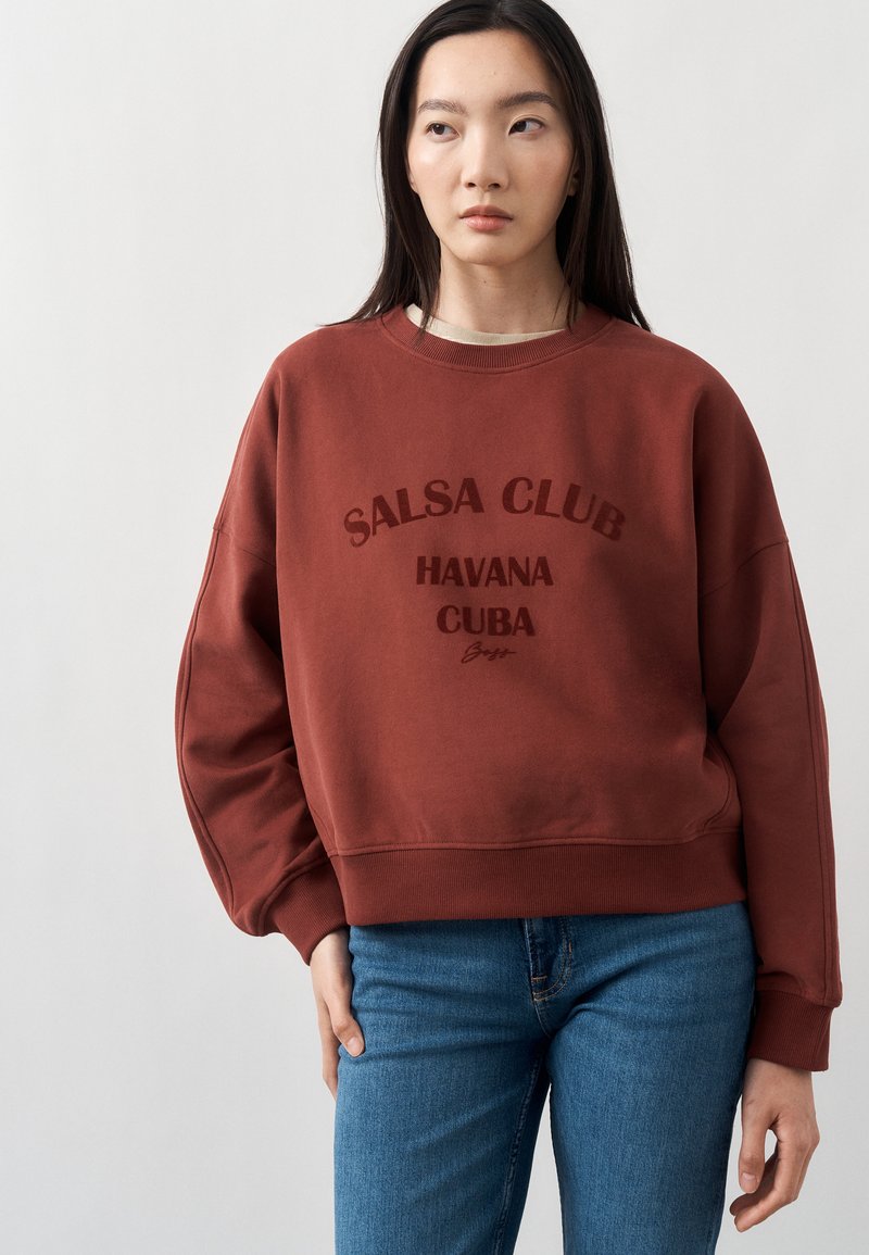 Femme aux longs cheveux foncés portant un sweat-shirt couleur rouille avec l’inscription « Salsa Club Havana Cuba » et un jean bleu sur un fond clair uni.