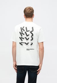 T-shirt branca de algodão com um design gráfico preto nas costas, apresentando letras e padrões geométricos. Gola redonda, mangas curtas, corte descontraído.