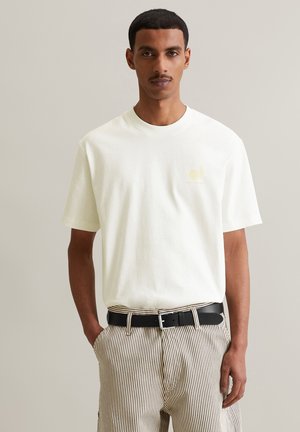 White cotton t-shirt with a round neckline and short sleeves, featuring a subtle yellow print logo on the chest. Worn with striped pants.