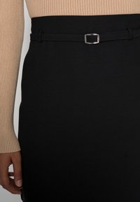 Zwarte rok met een gladde textuur, voorzien van een dunne riem met een vierkante zilveren gesp. Naast de ribgebreide beige top zichtbaar bij de manchetten.