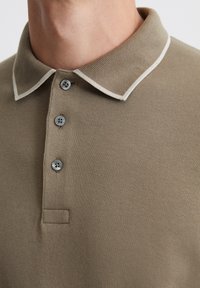 Hellbraunes Poloshirt aus strukturiertem Stoff mit einem kontrastierenden weißen Kragen, drei grauen Knöpfen und einem kleinen Stoffpatch in der Nähe der Knöpfe.