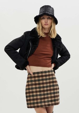 Giacca nera foderata in pelliccia sintetica, maglione corto a coste marrone e una mini gonna a quadri beige con accenti marroni e verdi. Un cappello a secchiello nero completa il look.