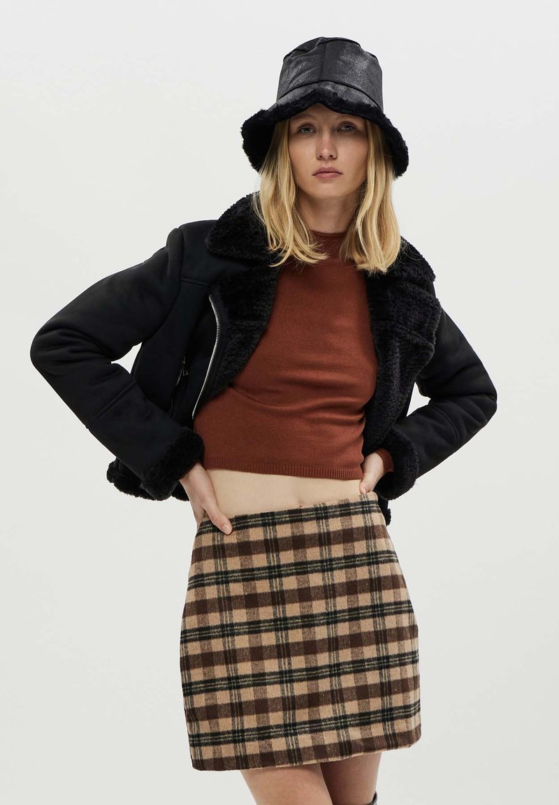 Giacca nera foderata in pelliccia sintetica, maglione corto a coste marrone e una mini gonna a quadri beige con accenti marroni e verdi. Un cappello a secchiello nero completa il look.