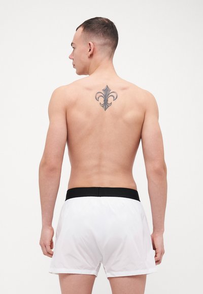 Shorts de bain blancs avec une taille noire, vus de dos. Inclut un tatouage noir de fleur de lys sur le haut du dos. Texture de tissu lisse.