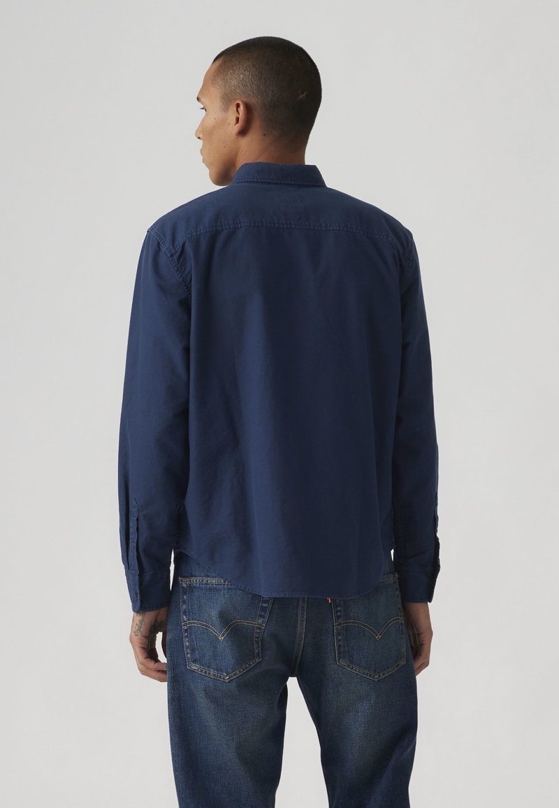 Hombre de espaldas vestido con una camisa azul marino de manga larga y vaqueros azules sobre un fondo claro y liso.