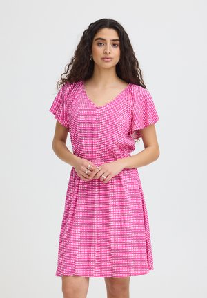 Kobieta z długimi kręconymi włosami, ubrana w różową sukienkę do kolan z białymi wzorami serduszek i zwiewnymi rękawami, stoi na jednolitym tle.