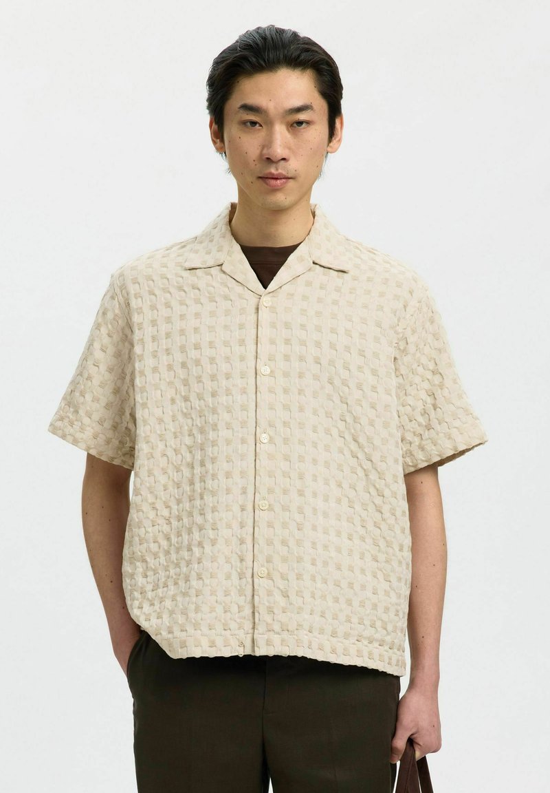 Jeune homme portant une chemise beige texturée à manches courtes boutonnée par-dessus un sous-vêtement marron, debout devant un fond blanc uni.