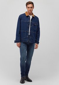 Denim jacket in dark blue with a tan collar, featuring front pockets, button closure, and long sleeves. Paired with blue jeans and black boots.