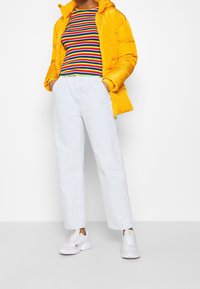 Gul pufferjacka, flerfärgad randig skjorta, ljusblå vida jeans och vita sneakers; släta texturer med avslappnad passform.