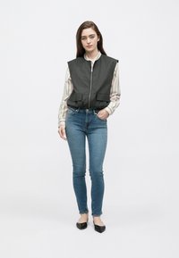 Colete cinzento escuro sem mangas com fecho de correr frontal e dois bolsos frontais, usado sobre uma camisa branca de mangas compridas com riscas e jeans azuis. Sapatos planos pretos.