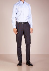 Světlomodrá košile s límečkem do špičky, doplněná hnědými károvanými kalhotami s modrými a červenými akcenty a černými šněrovacími botami.