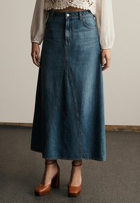 Denim maxi skirt, dark blue, with an A-line silhouette. Features front seams and a buttoned waistband. Paired with brown heeled shoes.