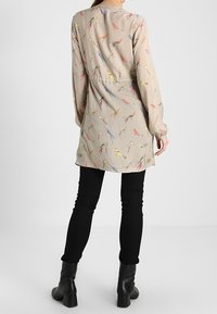 Beige Tunika mit bunten Vogelmustern; leichter Stoff; lange Ärmel; kombiniert mit schwarzen Hosen und Ankle Boots; Rückansicht.