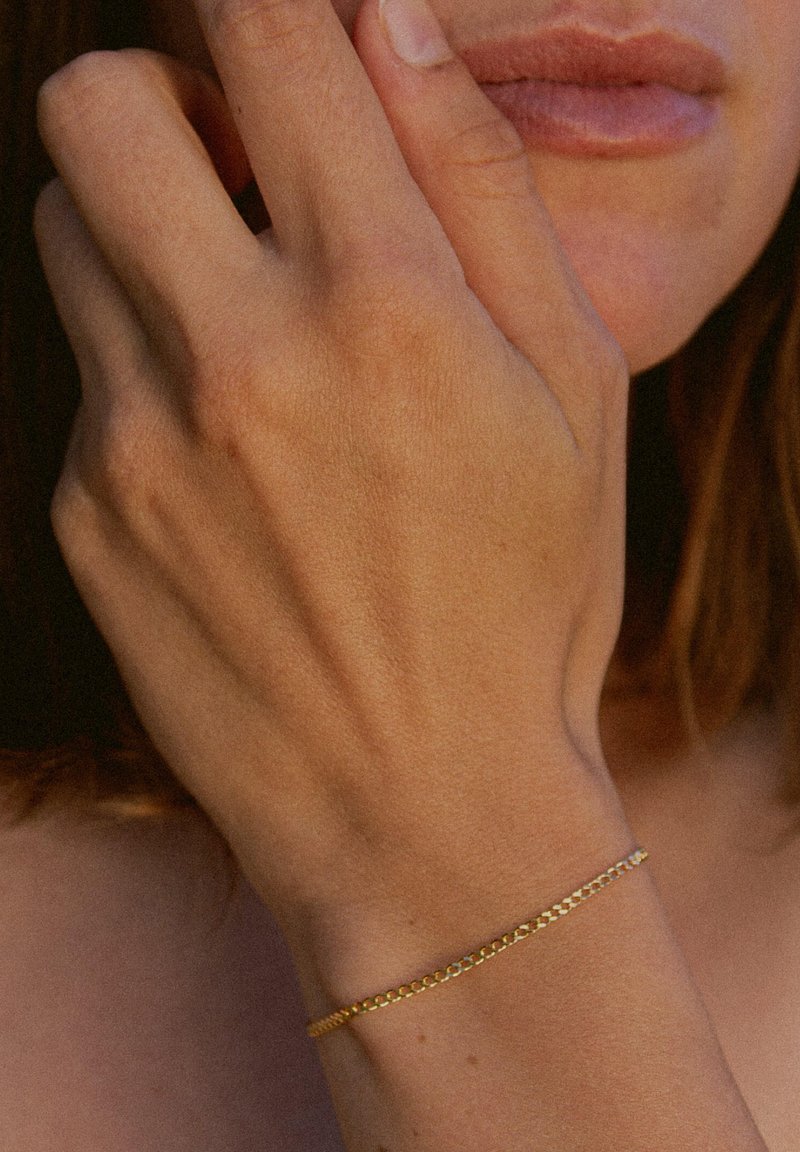 Goldenes Kettenarmband an einem Handgelenk, fein gestaltet mit ineinandergreifenden Gliedern, glatter Textur und minimalistischem Stil. Keine zusätzlichen Verzierungen.