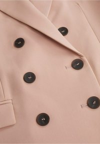 Blazer croisé en tissu rose doux, avec des boutons noirs. Les détails remarquables incluent une texture lisse et des revers structurés.