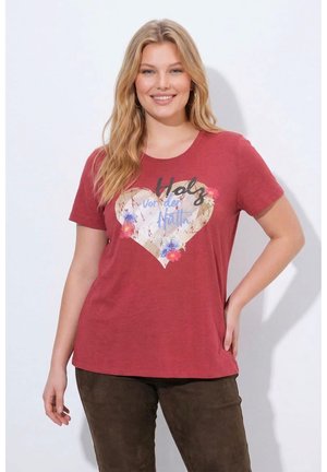 Lächelnde Frau mit langen blonden Haaren trägt ein rotes T-Shirt mit Herz- und Blumenmotiv und dunkle Hose, steht mit einer Hand in der Hüfte.