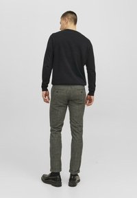 Un pull noir à col rond associé à un pantalon gris à motifs comportant des nuances de vert et de brun, porté avec des chaussures noires.