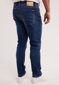 Bărbat purtând blugi slim-fit, albaștri închiși, cu buzunare în spate și adidași albi, stând pe un fundal simplu, deschis la culoare.