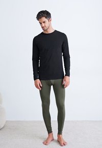 Svart langermet skjorte kombinert med olivengrønne tights, tettsittende design, glatt tekstur, brukt barfot på et lyst teppe.