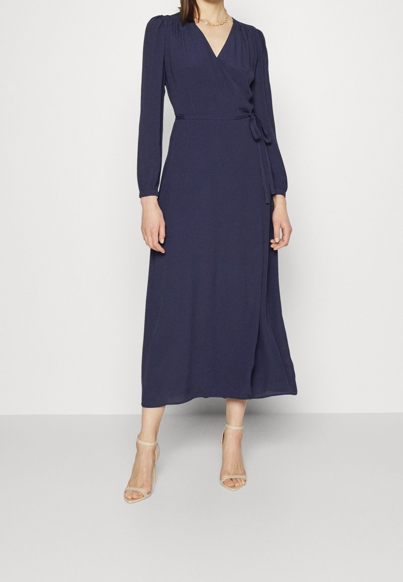 Femme portant une robe portefeuille bleu marine à manches longues avec une ceinture à la taille et des sandales à talons beiges, debout devant un fond uni.